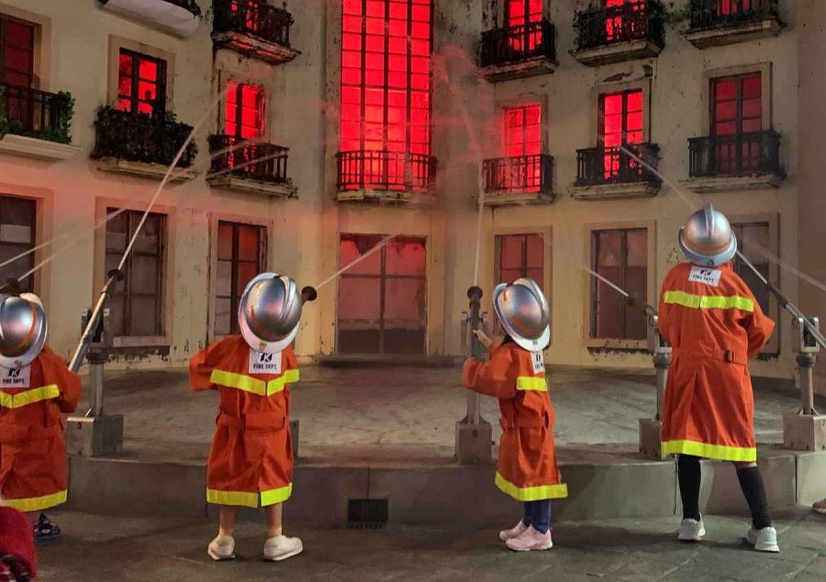 Kids in firefighter uniforms aiming hoses at a mock burning building